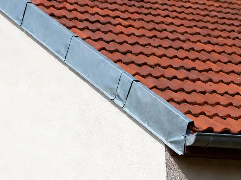 Bande de rive - Couverture Travaux Gros plan d'un toit de maison en pente avec des tuiles en terre cuite rouge et une bande de rive en métal le long du bord où le toit rencontre le mur extérieur blanc.