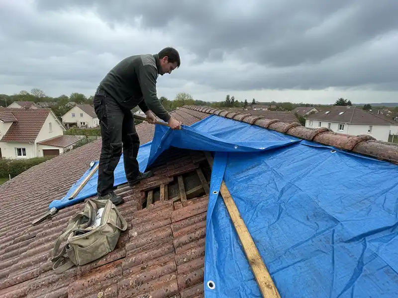 Infiltration eau toiture - Couverture Travaux Un couvreur se tient sur un toit de tuiles en pente, fixant une bâche bleue sur les tuiles endommagées pour empêcher l'infiltration d'eau dans la toiture à l'aide de planches de bois par une journée nuageuse. Un sac à outils se trouve à côté d'eux et des maisons sont visibles à l'arrière-plan.